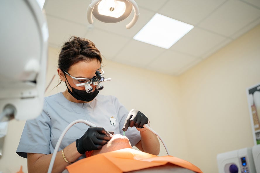 Female dentist in binocular glasses treats a man in exam chair